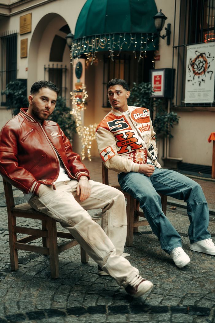Two fashionable young men in stylish jackets sitting outdoors in a vibrant city setting.