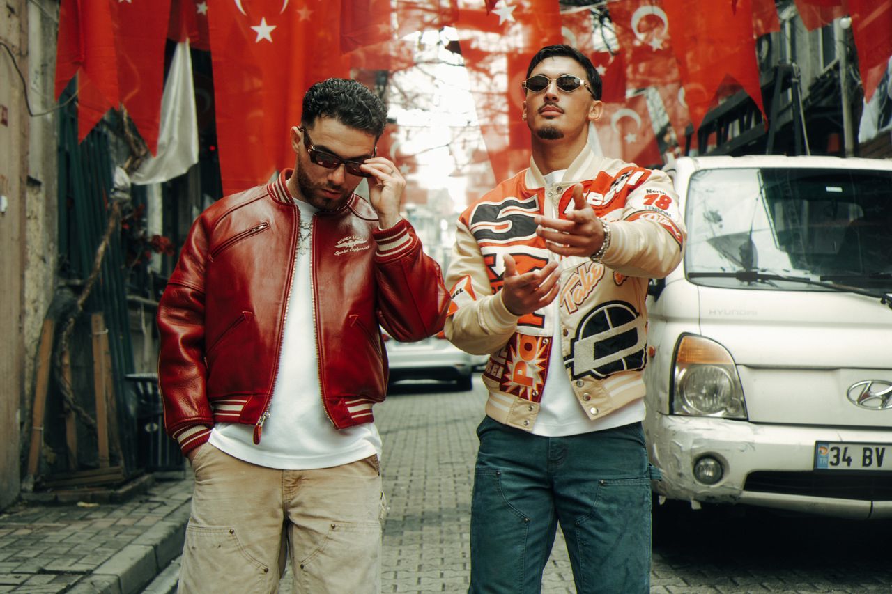 Two fashionable men on city street with red flags hanging, showcasing urban style.